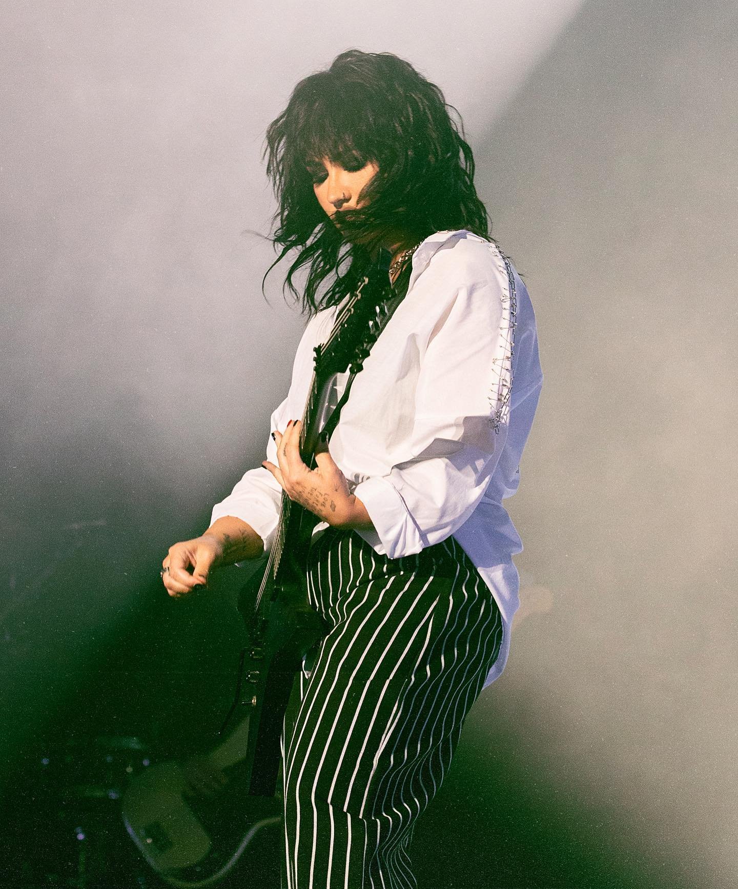 A person with dark hair performing on stage, wearing a white shirt and striped pants, with stage lighting and a musical instrument.