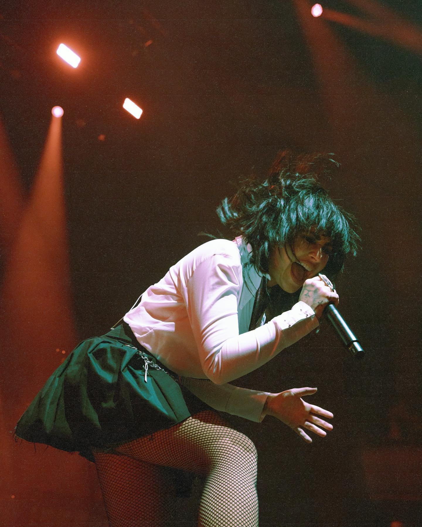 A woman sings on stage wearing a white shirt, black skirt, and fishnet stockings, with a microphone in hand and stage lighting.