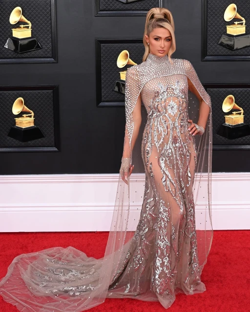 Paris Hilton on a red carpet wearing a shimmering gown with a high neckline and a long train posing in front of a dark wall.