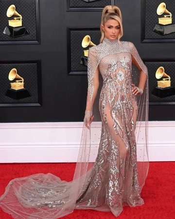 Paris Hilton on a red carpet wearing a shimmering gown with a high neckline and a long train posing in front of a dark wall.