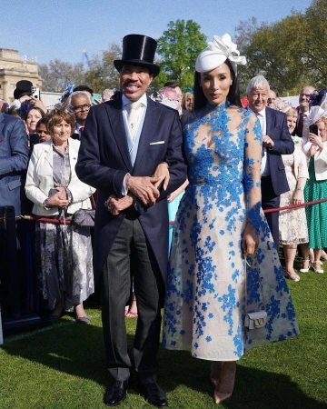 Couple in formal attire at an outdoor event: a man in a top hat and suit with a woman in a blue and white dress and fascinator hat.