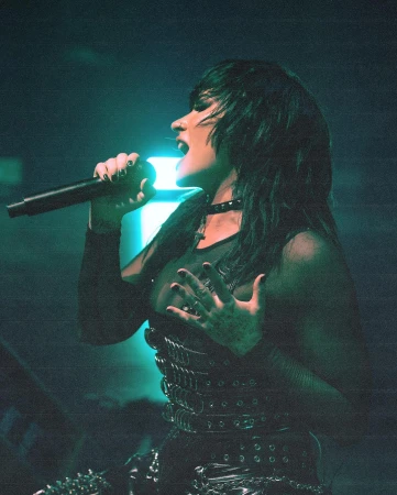 Female singer performs on stage with microphone, wearing a studded top and choker, with green stage lighting creating a dramatic effect.