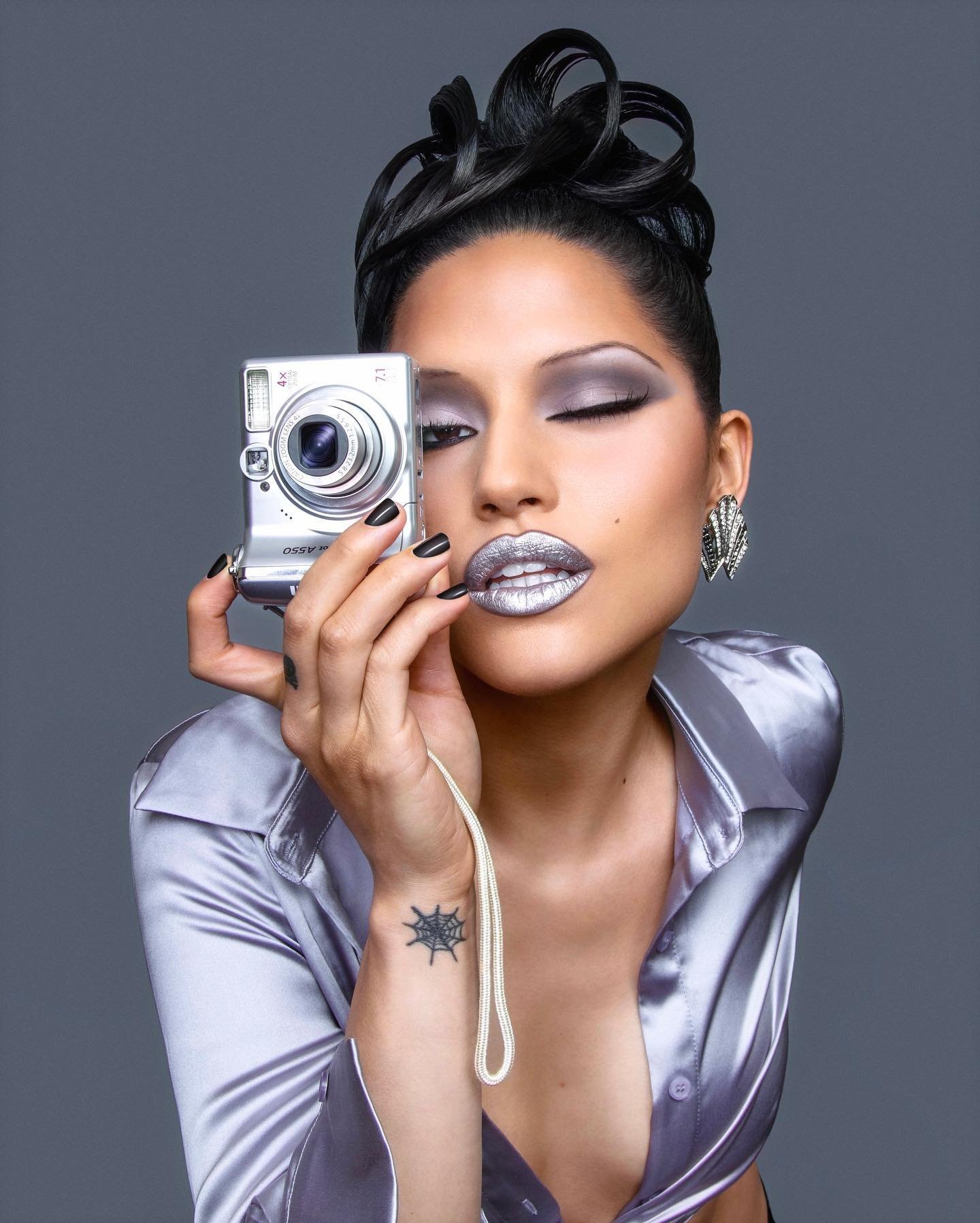 Headshot of a woman holding a silver camera to her face, wearing silver eyeshadow, silver lipstick, and a satin button-down shirt in a studio.