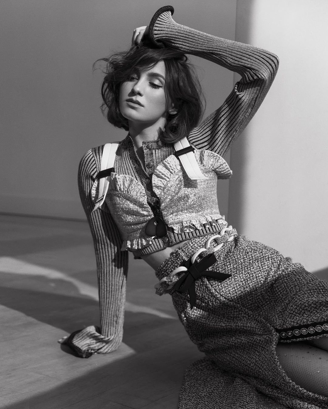 Monochrome portrait of a woman with wavy bob, wearing a textured crop top and skirt, posing on a floor in a minimalistic studio setting.