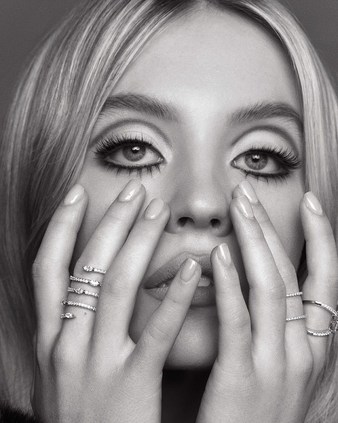 Monochrome close-up of a woman with fair skin and blonde hair with her hands near her face wearing diamond rings in a studio.