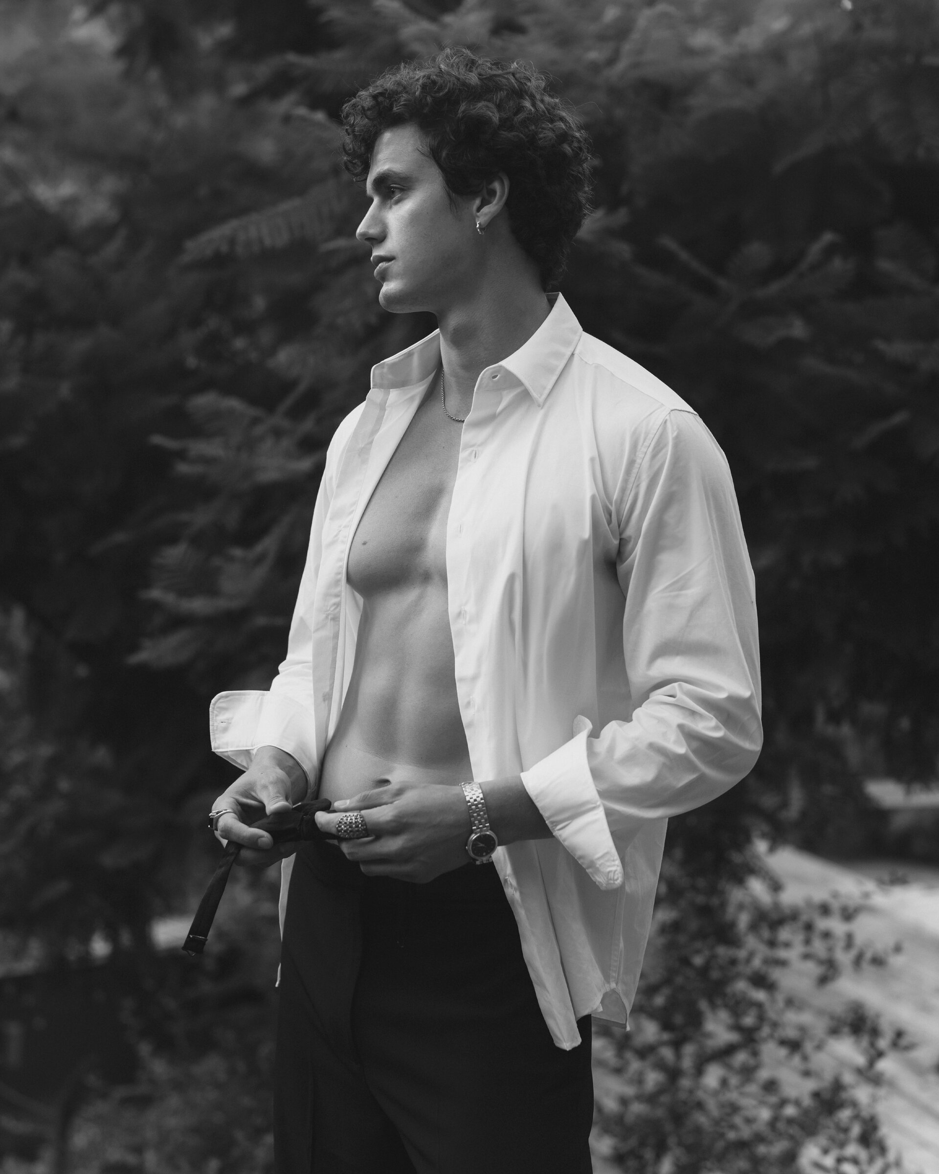 Monochrome portrait of a curly-haired man in an open white shirt and dark pants standing in a leafy green outdoor setting looking left.