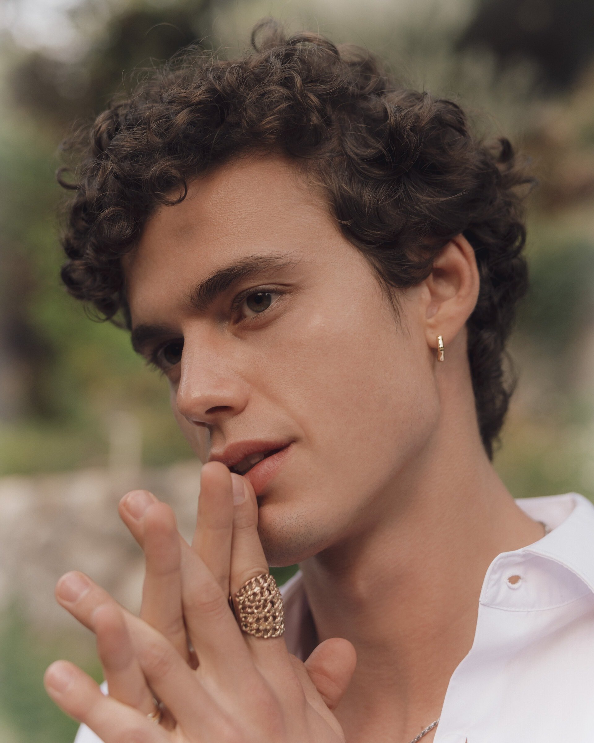 Close-up portrait of a curly-haired man with a gold ring, wearing a white shirt, with a blurred outdoor background. Fashion advertisement.