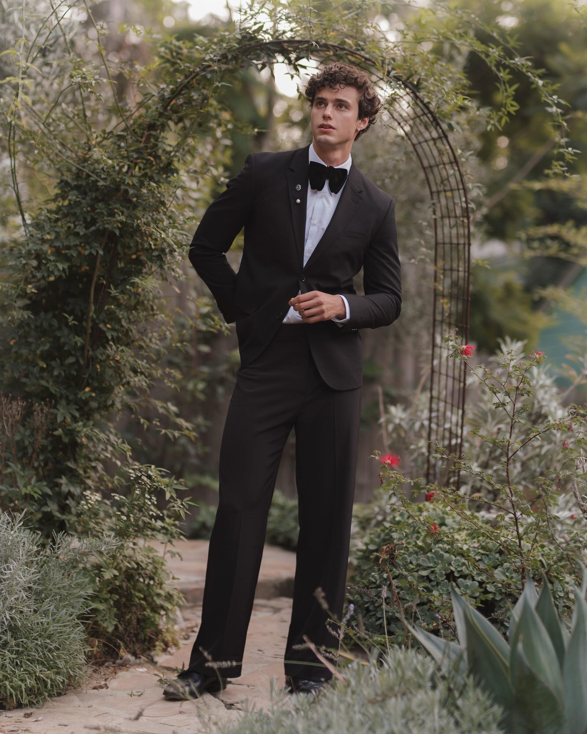 Full-length shot of a young adult male wearing a black suit and bow tie, posing in a garden setting with an archway behind him.