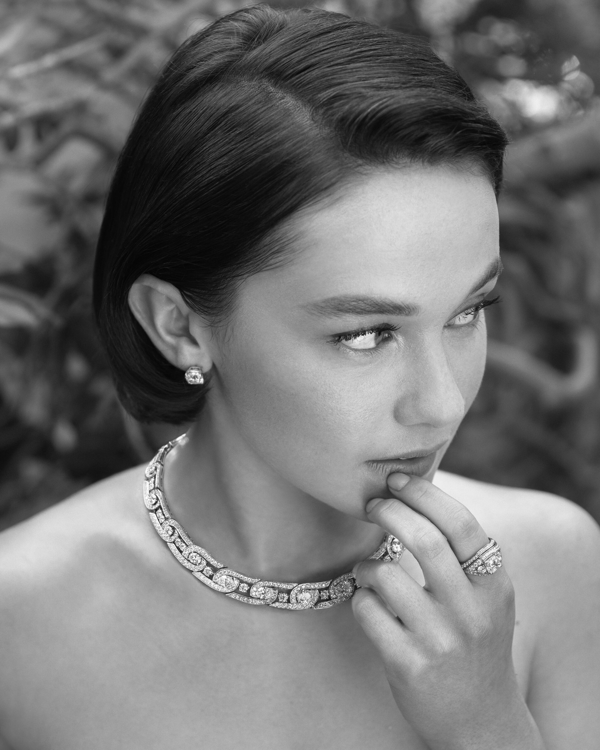 Black and white portrait of a young woman with dark hair wearing diamond jewelry including a necklace, earrings, and rings outdoors.