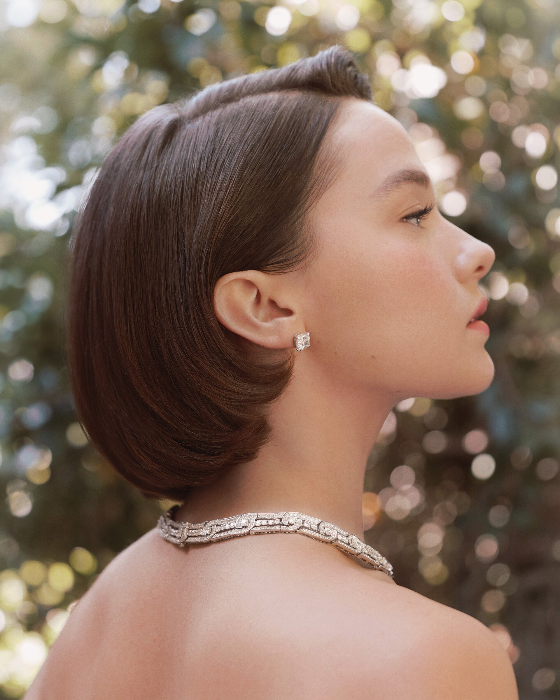 Profile portrait of an adult woman with short brown hair and a diamond necklace and earrings, set against a blurred background of green foliage.