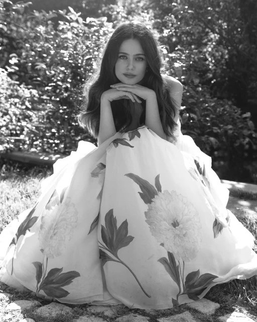 Monochrome portrait of a woman in a floral dress, sitting outdoors with hands resting on her chin, framed by lush foliage.