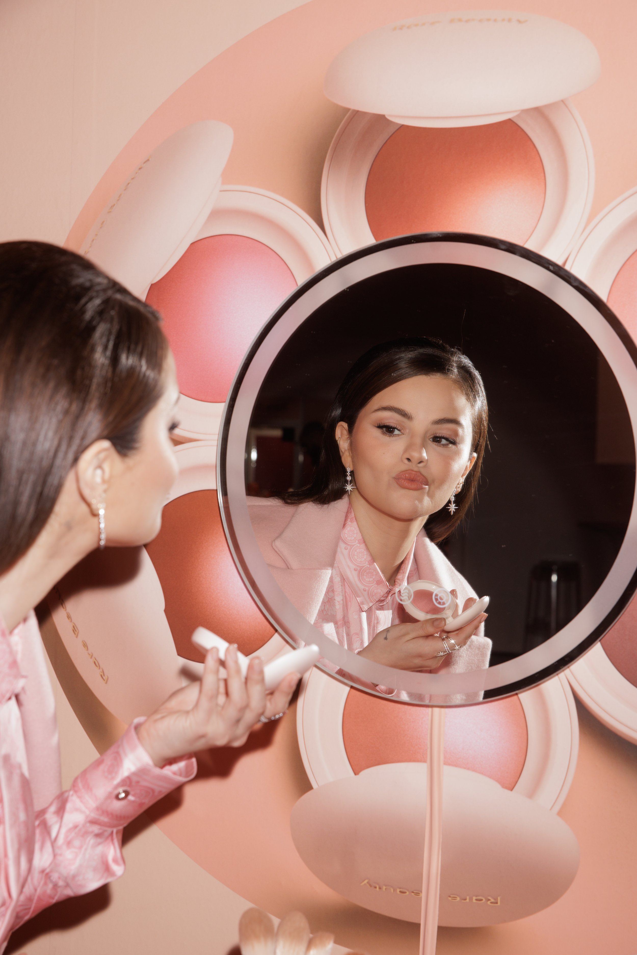 Selena Gomez gazes into a lit mirror while holding a cosmetic product in a beauty campaign, wearing a pink suit and earrings.