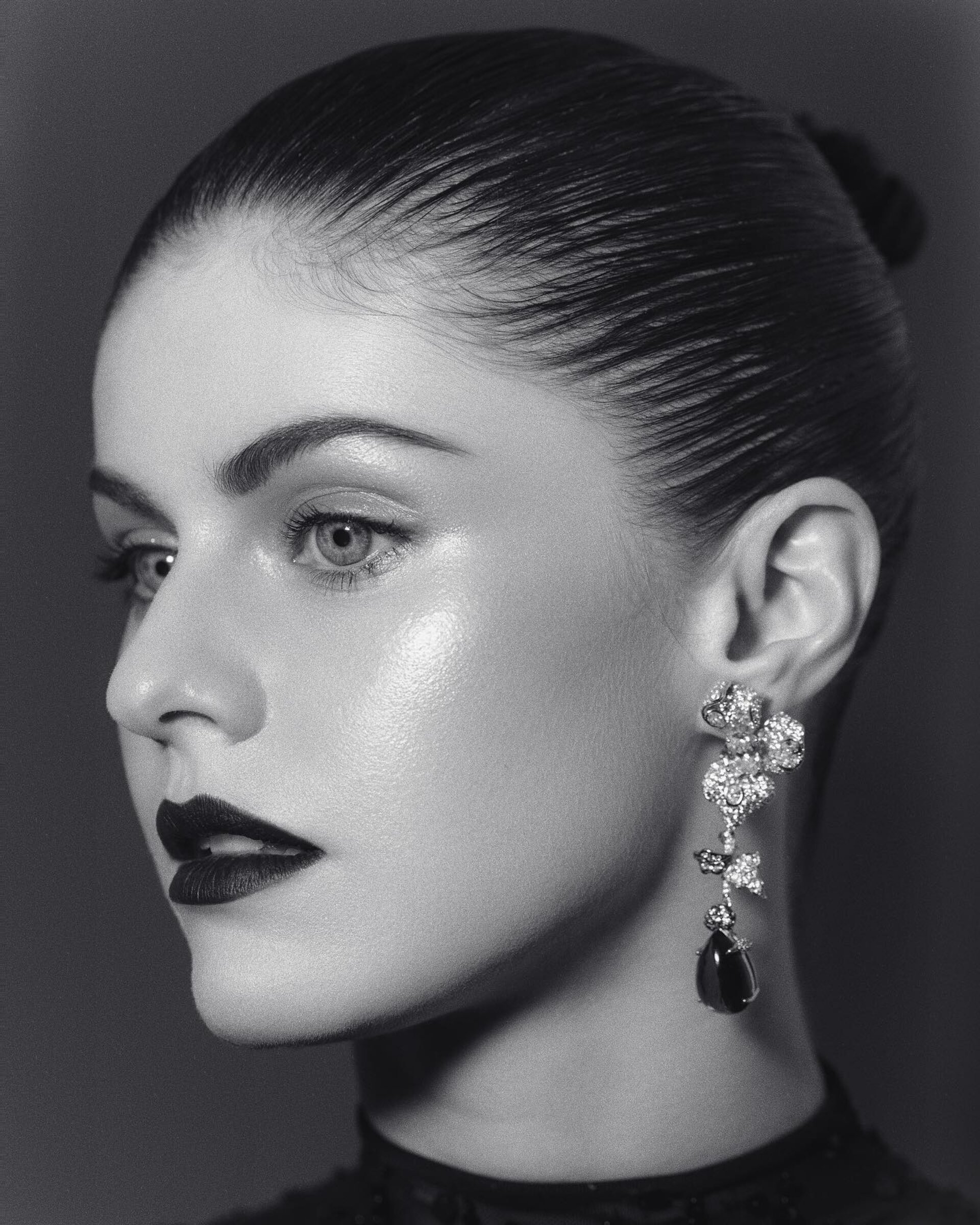 Monochromatic portrait of a woman with slicked-back hair, dramatic makeup, and floral drop earrings against a gray studio backdrop.