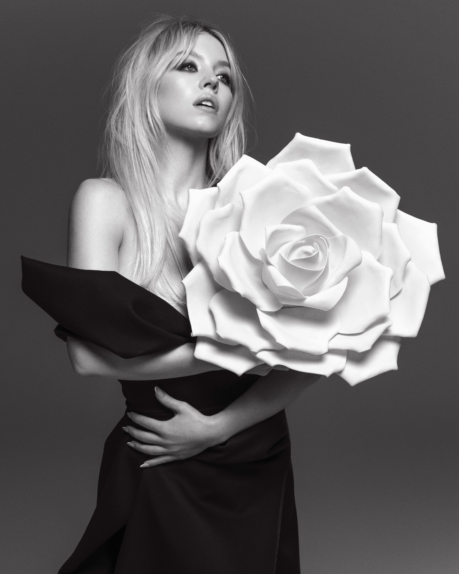 Black and white portrait of a blonde woman in a black dress, holding a large white paper flower, posing in front of a solid grey background.