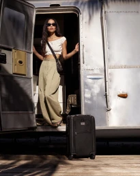 Zoe Kravitz wearing sunglasses and high-waisted trousers standing in an Airstream doorway with a black suitcase
