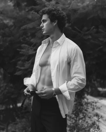 Portrait of a man with curly hair wearing an open white shirt and dark trousers against foliage