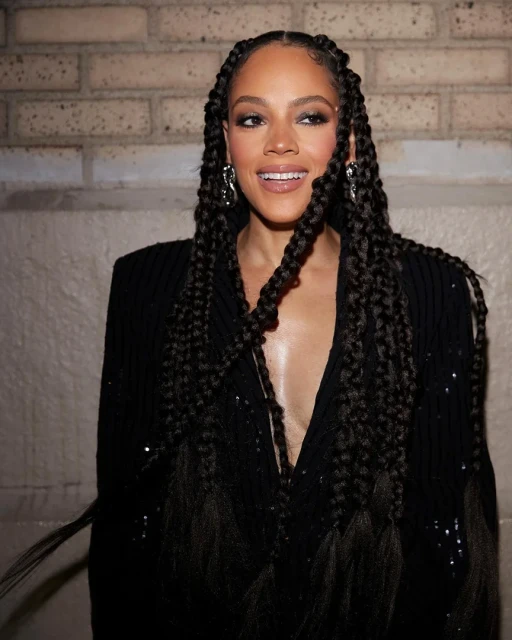 Close-up of a woman with braided hair wearing a sparkling black jacket, smiling in front of a neutral brick background.