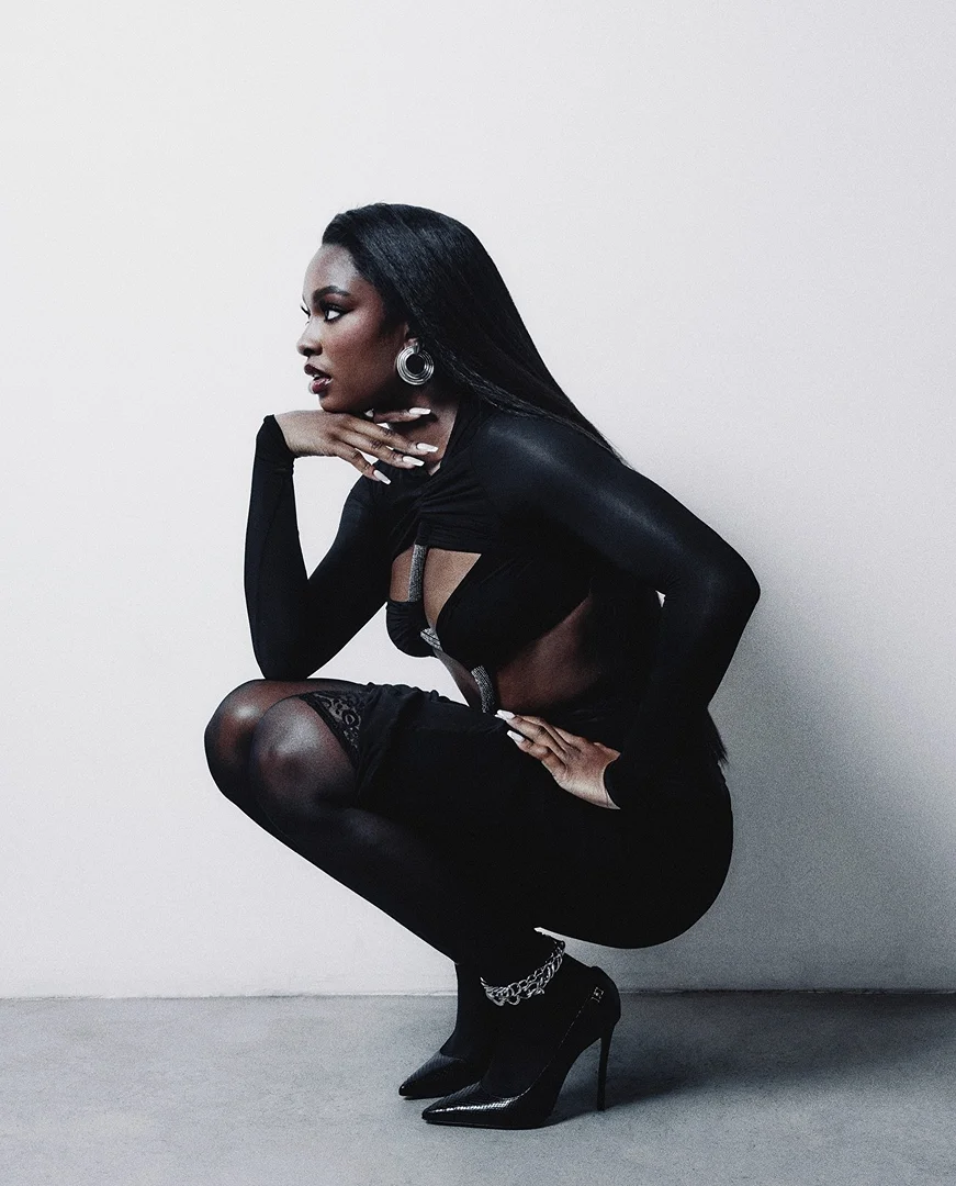 Profile shot of a Black woman with long straight hair, wearing a black outfit and high heels, crouching in a photo studio.