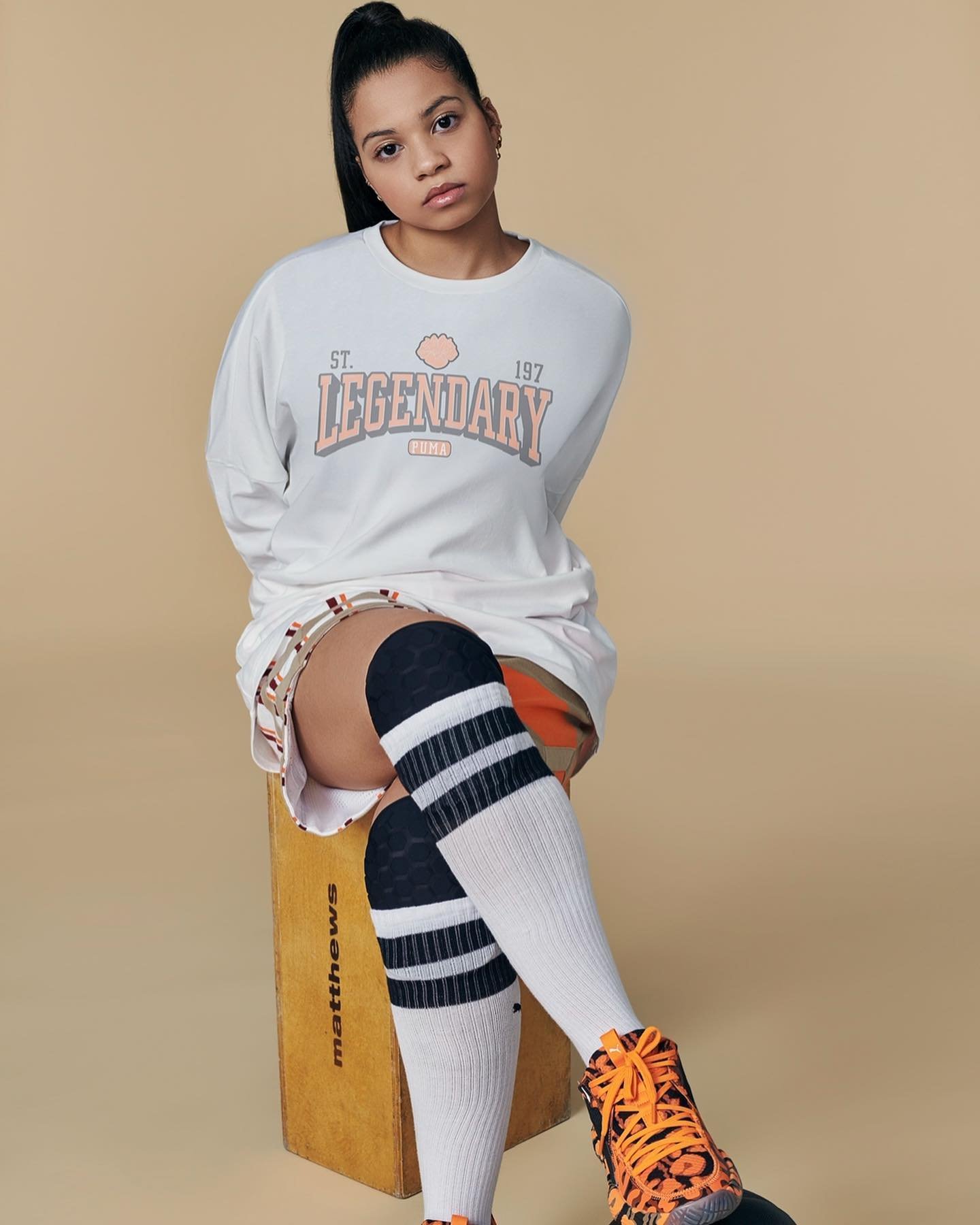 Woman in a white long sleeve shirt with a graphic, seated on a wooden box with orange basketball and sneakers in a studio setting.