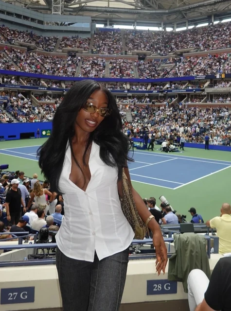 Woman with long black hair at a tennis match wearing a white blouse and jeans in a stadium filled with spectators.