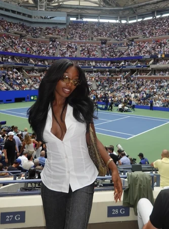 Woman with long black hair at a tennis match wearing a white blouse and jeans in a stadium filled with spectators.