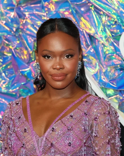 Close-up portrait of a smiling Black woman with long dark hair wearing chandelier earrings and a pink beaded dress against a holographic background.