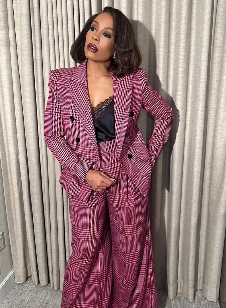 Woman in pink plaid suit poses indoors against neutral curtain. She wears dark lipstick, and her hair is styled in a wavy bob.