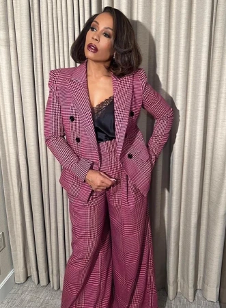 Woman in pink plaid suit poses indoors against neutral curtain. She wears dark lipstick, and her hair is styled in a wavy bob.