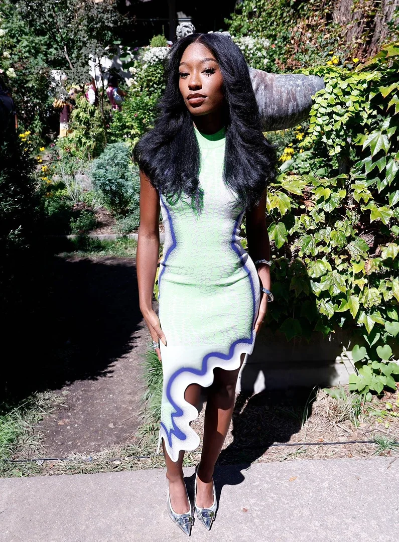 A dark-skinned woman poses outdoors wearing a green, white, and purple dress and silver heels near greenery and a concrete path.
