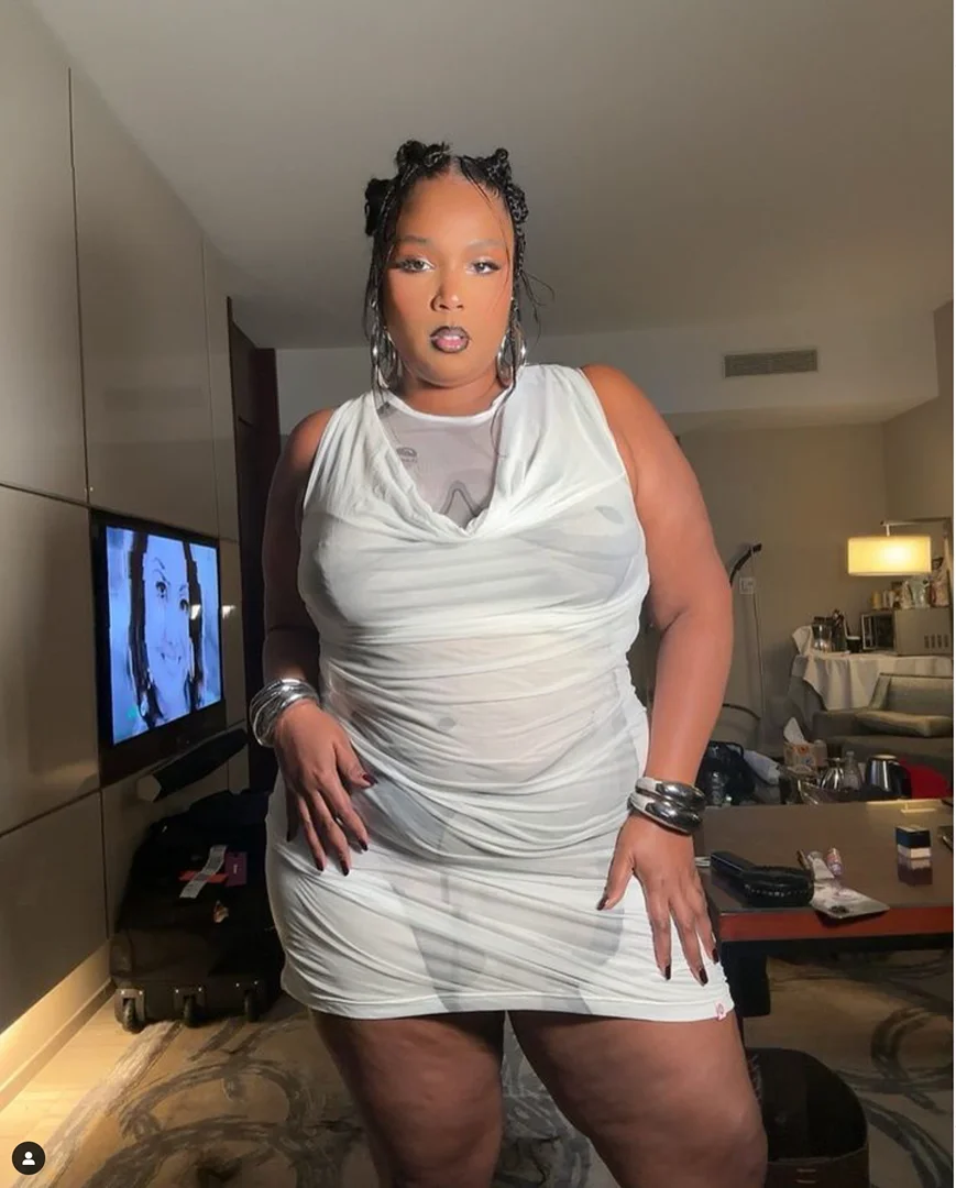 A woman with braided hair poses in a sleeveless white dress and metal bracelets in an interior setting with a TV and furniture.