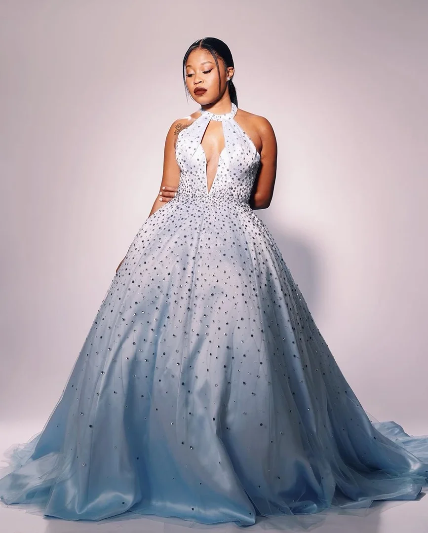 Woman wearing a pale blue gown with embellished detail poses in front of a gradient grey backdrop in a fashion editorial shot.