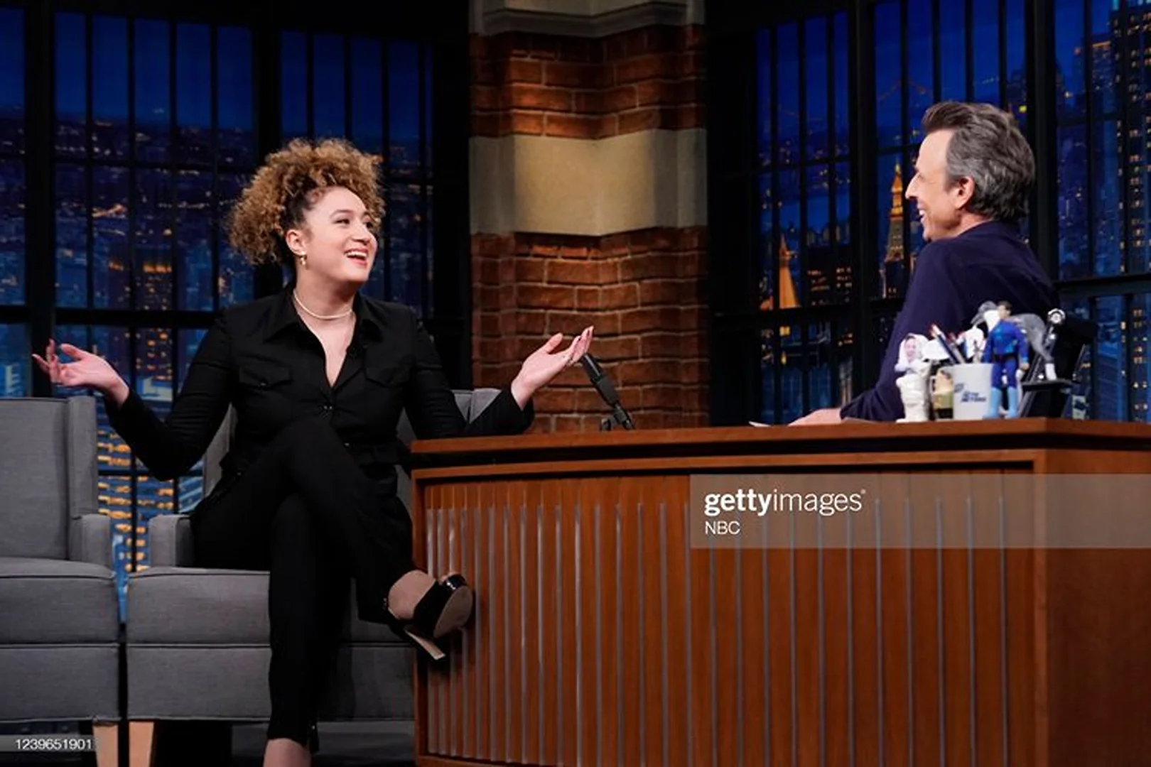 Two adults on a late-night talk show set, a woman in black sits on a gray couch while a man sits at a desk against a brick wall.