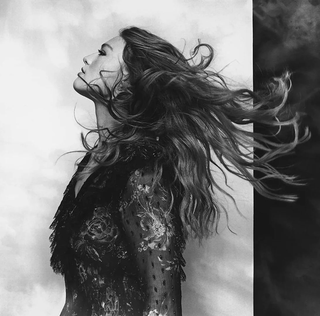 Black and white portrait of a woman with windswept wavy hair, wearing a floral top, against a mottled gray and black background.