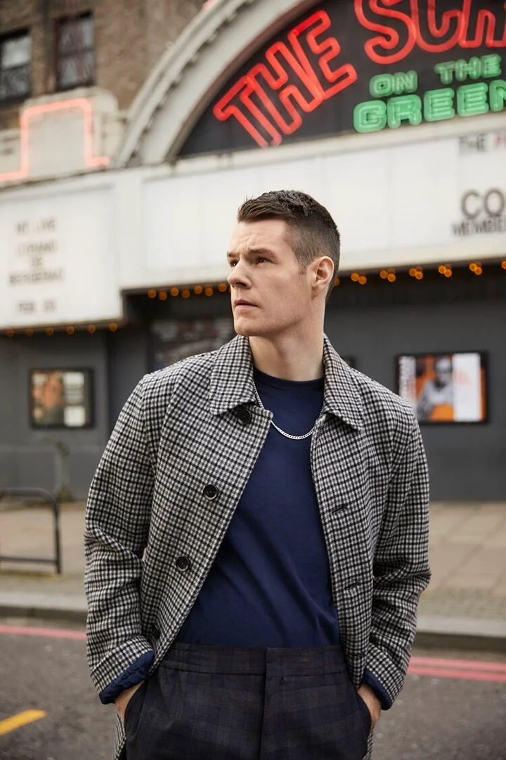 Man in checkered overcoat stands in front of the neon-lit entrance to The Screen on the Green cinema on a cloudy day.