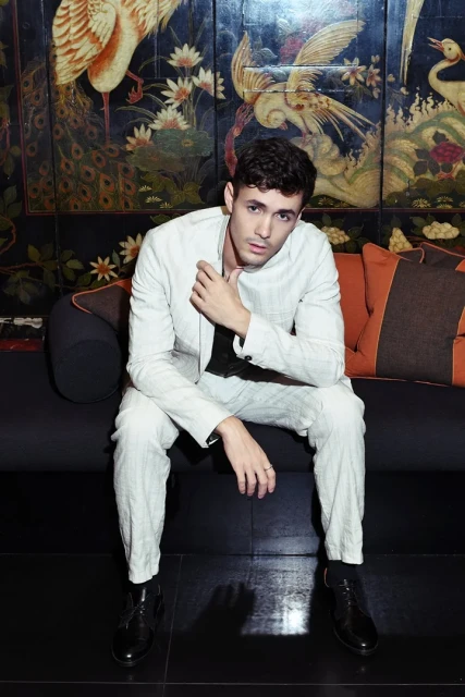 A young man in a light-colored suit poses seated in front of an ornate, dark-toned wall featuring birds and floral designs.