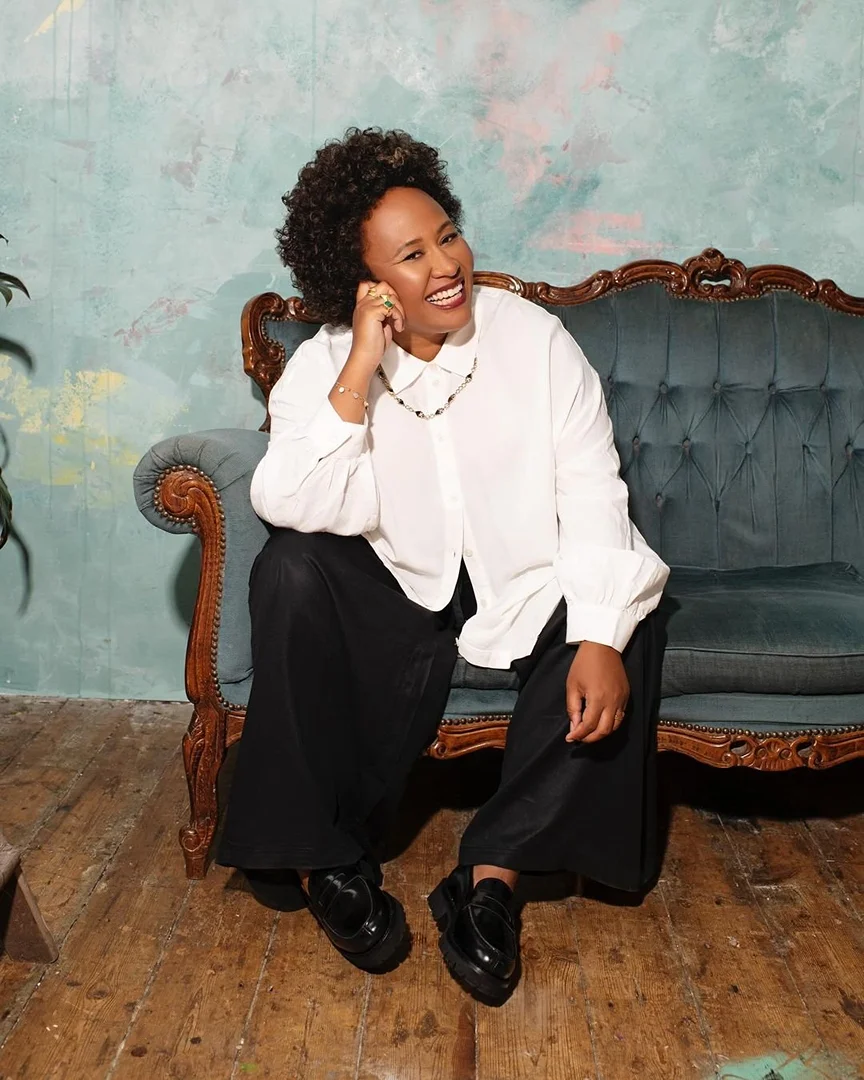 A smiling Black woman with short curly hair sits on a vintage couch wearing a white blouse and black pants in a casual indoor portrait.