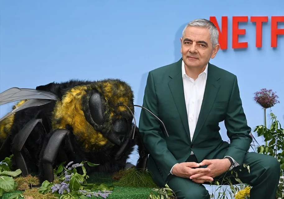 Rowan Atkinson poses next to a large model bee; he wears a green suit and white shirt against a light blue background with Netflix logo.