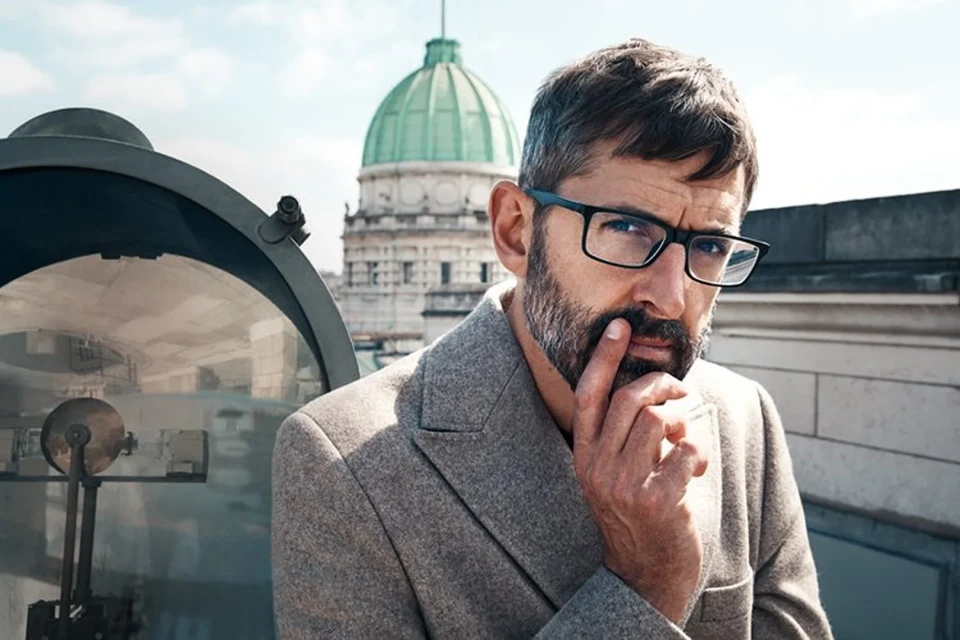 Adult male with glasses and beard stands on rooftop with city building dome and cloudy sky in background, wearing grey coat.
