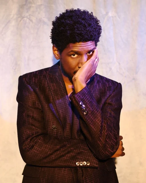 Portrait of a Black man in a suit with hand on face, posing against a textured backdrop with soft lighting, showing fashion and style.