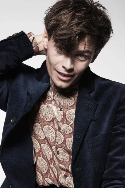 Portrait of a young adult male with dark hair, wearing a patterned shirt and corduroy jacket, posing indoors against a white backdrop.