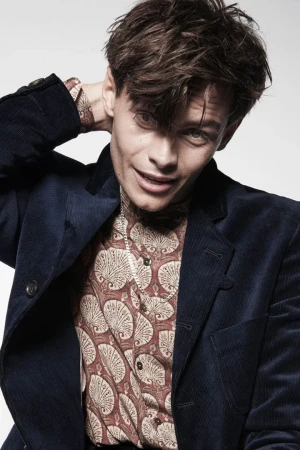 Young man in a navy corduroy jacket and patterned shirt with hand behind head against a plain light background
