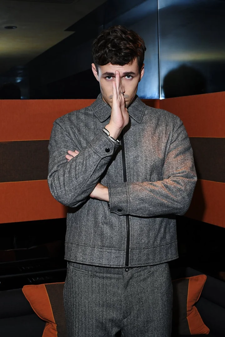 Portrait of an adult man wearing a grey tweed suit with his hands partially covering his face in an indoor setting with warm tones.