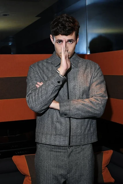 Portrait of an adult man wearing a grey tweed suit with his hands partially covering his face in an indoor setting with warm tones.
