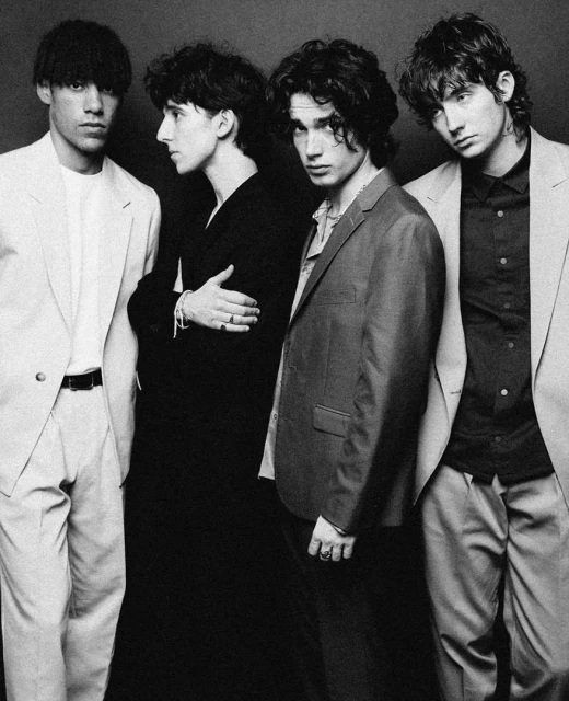 Four young adult men in suits pose indoors against a dark backdrop, in black and white. Three stand facing forward; one looks in profile.