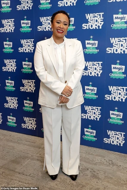 Stylish adult female in white suit at 'West Side Story' event, smiling, against a branded blue backdrop, captured in a full shot.