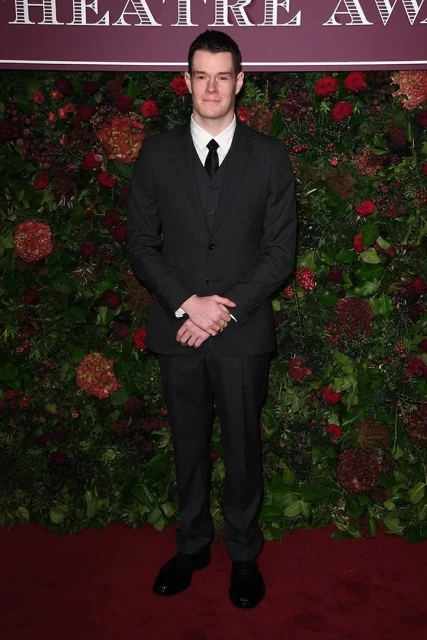 Full-length shot of a man in a dark suit with a floral backdrop at an event. He is wearing a black suit, vest, and tie.
