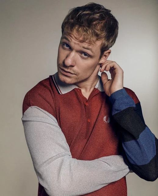 Close-up portrait of an adult male with blond hair wearing a red, gray, and blue polo shirt against a plain background.