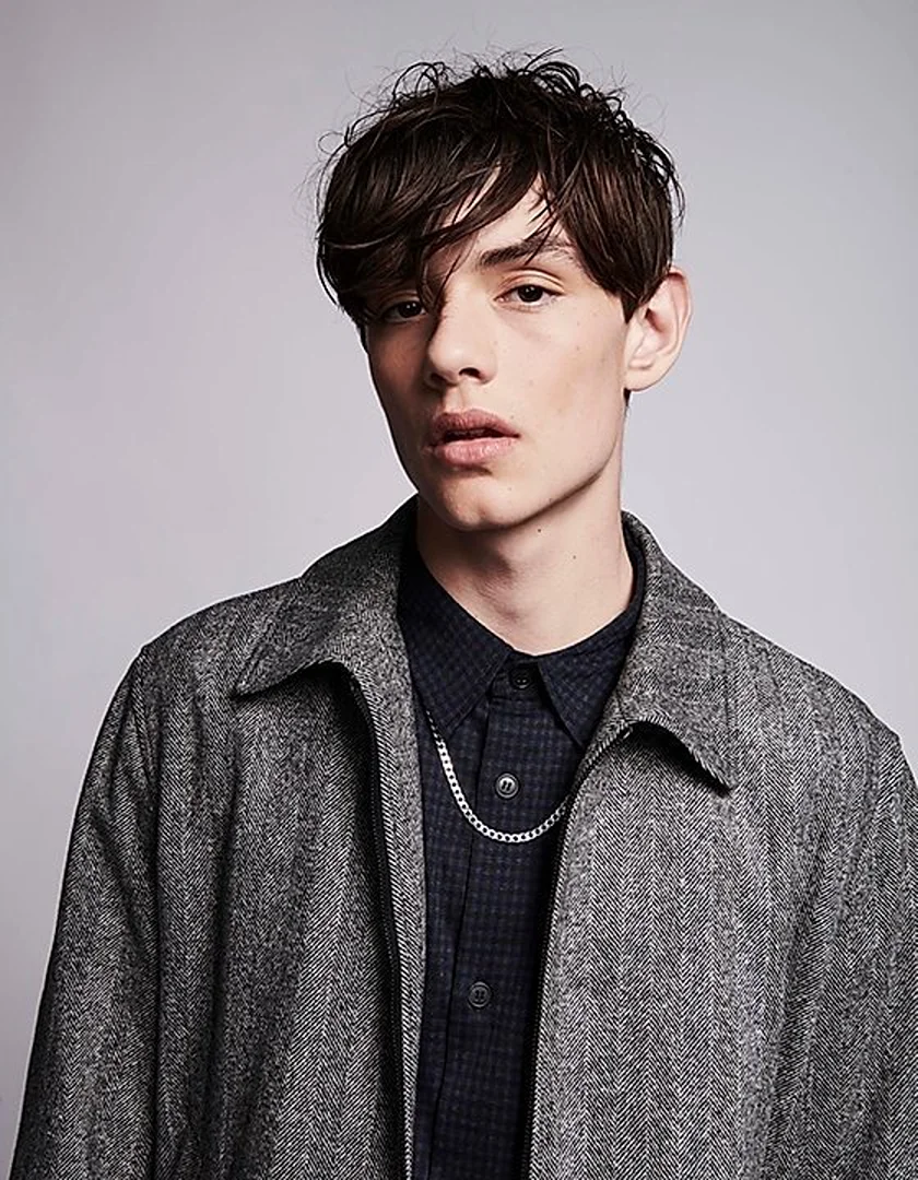 Portrait of a young adult man wearing a grey jacket and collared shirt with a silver chain necklace in a neutral studio setting.