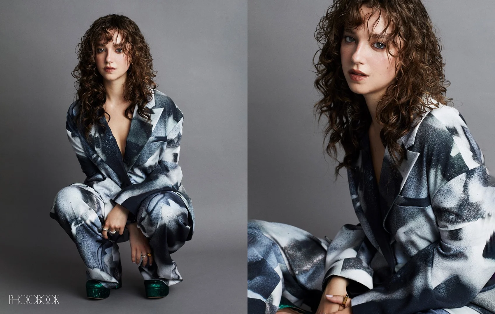 Young woman in a patterned suit poses in a photo studio with a grey backdrop. She has curly brown hair and is wearing green shoes.