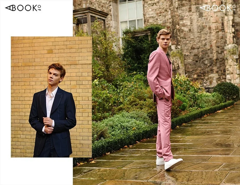 A young man in a pink suit stands on a stone path surrounded by greenery, with a smaller inset image of the same man in a navy suit.
